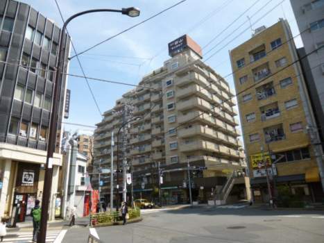 朝日白山マンションを交差点側から見上げた構図で、地上13階建の構造が段状に迫り上がる外観を捉えています。外壁はベージュ系タイルで仕上げられており、1階部分には店舗区画が設けられ、街路に開かれた構成です。電柱・電線が密集する都市型の環境も確認できます。朝日白山マンションは、昭和60年築、13階建てのマンションです。
