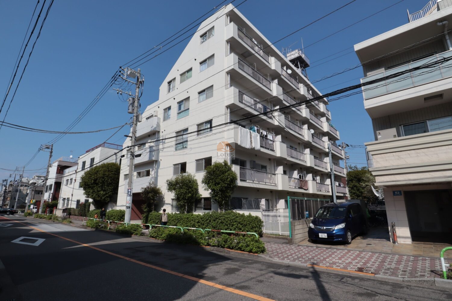 ヴェラハイツ西尾久の外観（南西側）を見上げています。白系外壁と連続するバルコニーが道路側に整い、歩道の生け垣が建物と通りをきれいに区切っています。角地側で視認性が高く、道案内がしやすい立地になっています。