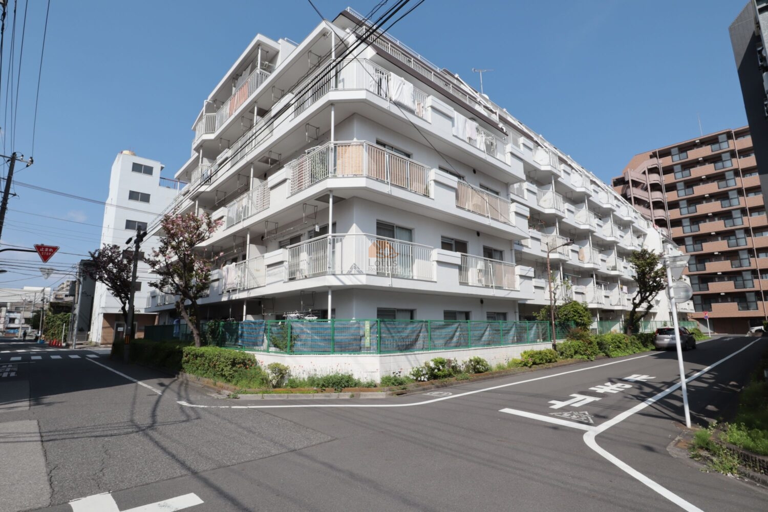 青い空のもと、白を基調とした爽やかな外観のマンションを、交差点の南西角から捉えた一枚。各フロアに配されたバルコニーが開放感を与えています。敷地周囲には植栽と緑色のフェンスが巡らされ、住居と道路を穏やかに区切っています。手前の道路には「止まれ」のペイントがあり、静かで落ち着いた住環境を想像させます。