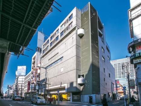 俳優座ビルの外観。六本木通りに面したSRC造の地上9階建で、コンクリート打ち放し風のファサードが1980年代の商業建築らしさを表しています。建物側面には円筒型の出窓が設けられ、視覚的なアクセントを形成。正面に広がる歩道と交差点は、視認性と来訪者の動線に寄与しています。