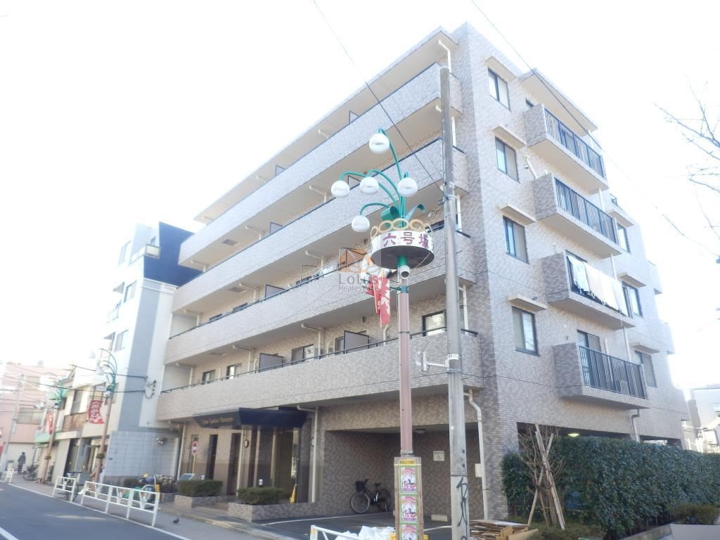 商店街の街路灯ポール(「六号通り」の表示)が立つ角地側からのライオンズガーデン幡ケ谷
の外観。タイル貼りの低〜中層ボリュームに、連続バルコニーと袖壁、コーナーに小さな出窓風の開口が見えます。1階はピロティ状で、自転車置き場と植栽が配置されています。電線・支柱・歩道柵を含む前面道路の様子も確認できます。