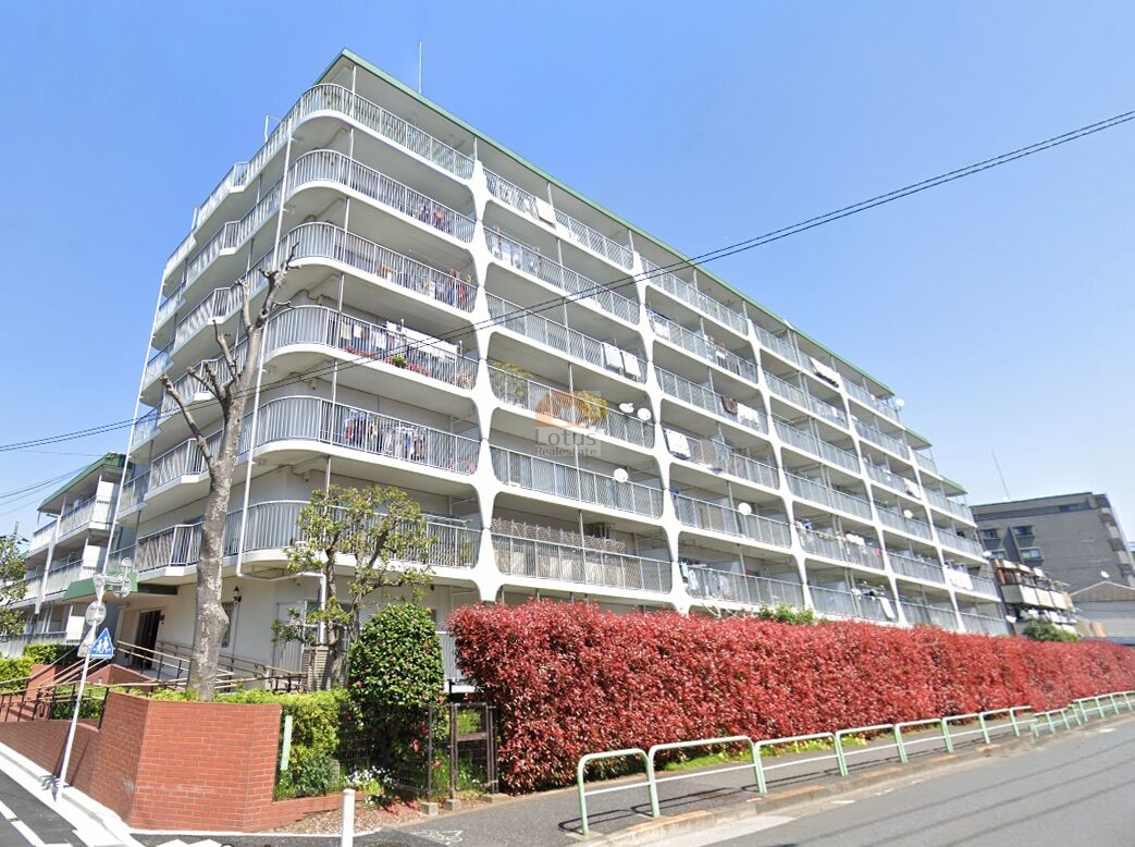 道路に面した住棟を見上げます。白い手すりのバルコニーが連続し、柱型の意匠が各階を上下に貫いています。歩道側には赤い生垣と街路樹、角にはレンガ調の擁壁が見えます。恒陽鷺宮マンションは、昭和53年築、7階建てのマンションです。