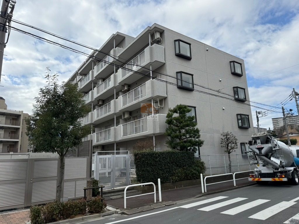 ユニーブル成増Ａ棟の外観。歩道と車道に面して、5階建ての住棟が見えます。金属手すりの連続バルコニー、角部の出窓、植栽帯、門扉とフェンスが確認できます。電柱や電線、道路標識も写り、周辺の街路の様子が分かります。ユニーブル成増Ａ棟は、平成2年築、5階建てのマンションです。