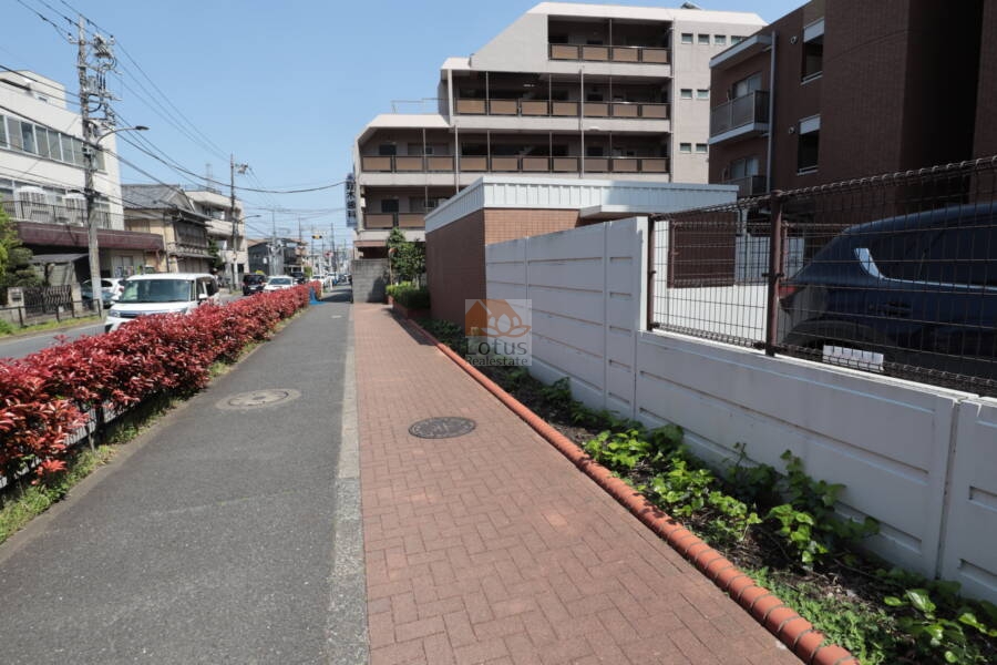 ブロッサムコートむさし野共用部1｜建物外周に面した歩道は、赤煉瓦調タイルと白い塀、植栽で整えられた印象的な外構。前面道路との境界も明確で、安全性と見映えの良さを兼ね備えています。