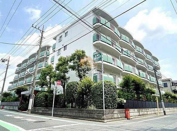 立石シティハイツ全体を斜めから撮影した外観で、白い外壁に半円形のバルコニーが連続して並んでいます。周囲には植栽がしっかり整えられ、道路に面した部分も緑で囲まれています。立石シティハイツは、昭和58年築、5階建てのマンションです。