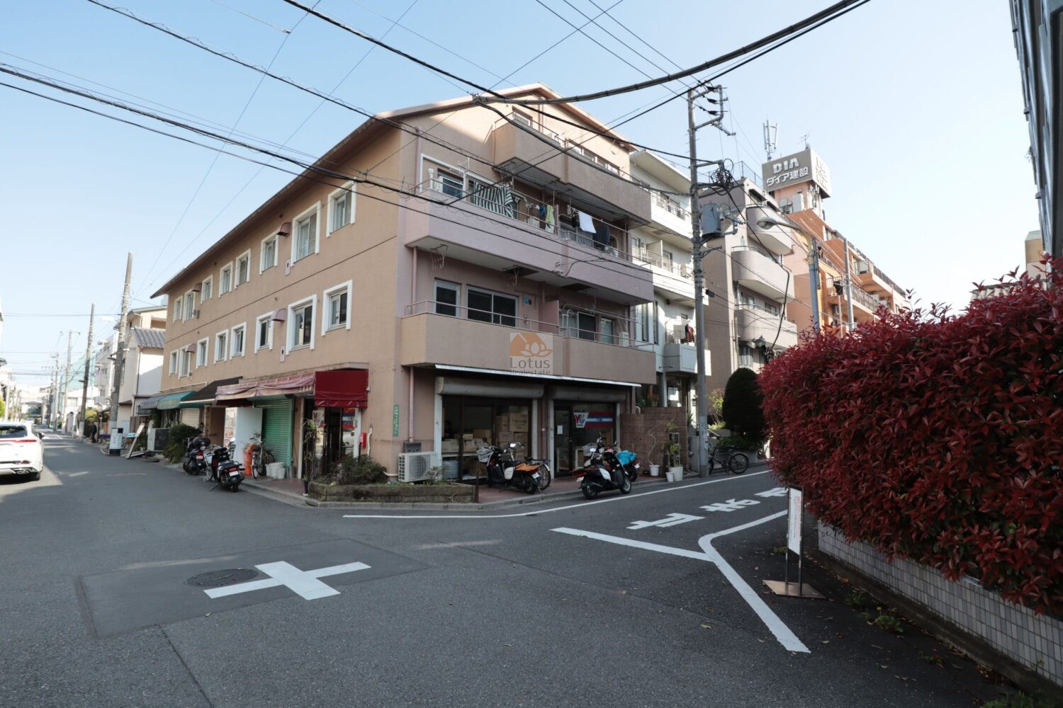 エルアルカサル尾久の外観（北西側）を見上げています。淡いブラウンの外壁が陽射しをやわらかく受けて、街並みに溶け込んでいます。1階には店舗が並び、角地ならではの開放感と生活のにぎわいを感じます。