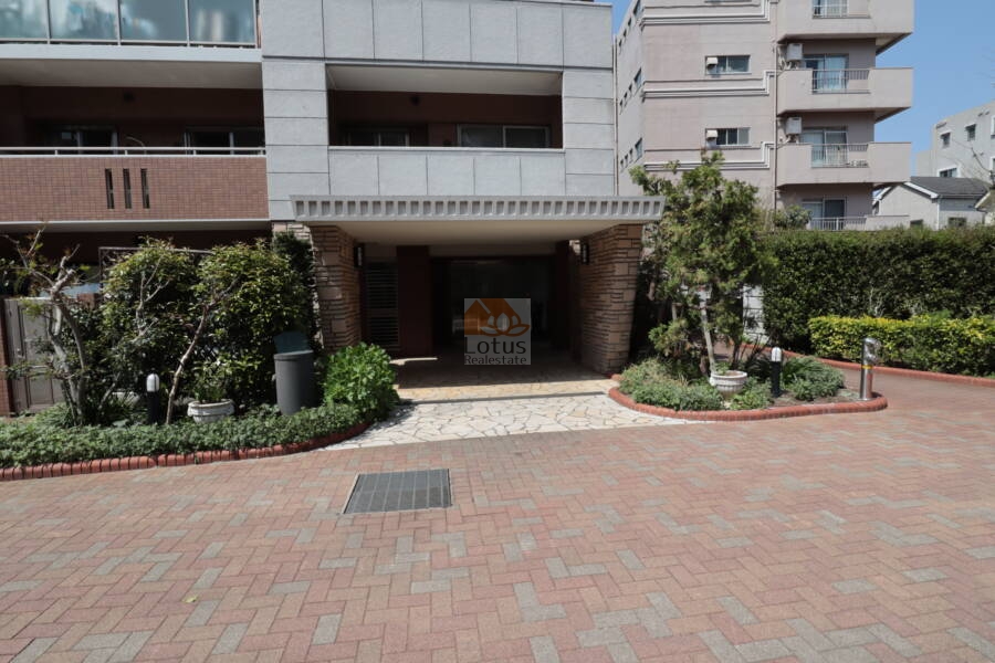ブロッサムコートむさし野ブライトウイングエントランス1｜ブロッサムコートむさし野のブライトウイングのエントランス。​庇が設けられ、エントランス前には植栽が配置されています。​アプローチはタイル敷きで、清潔感のある印象です。