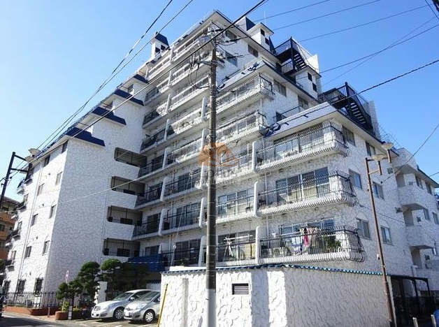 白い吹付仕上げの外壁に青い屋根と鉄柵のバルコニーが並び、南欧風の印象を与えます。道路に面したファサードは段状に構成され、上層階ほど後退するデザインで採光を確保しています。建物前面には駐車スペースと植栽が整えられ、周囲の街並みに明るい統一感を生んでいます。秀和小岩レジデンスは、昭和48年築、10階建てのマンションです。