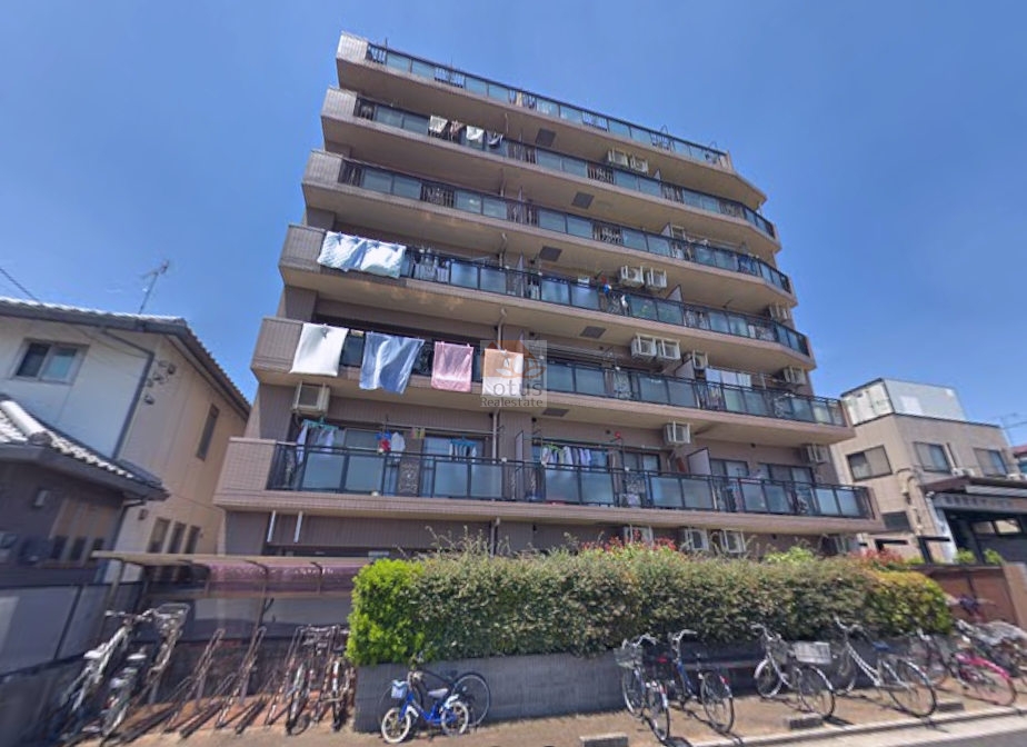 清潔感のあるブラウン系の外壁に、ガラスパネルのバルコニーがモダンな印象を与えるマンションです。写真から見えるように、各住戸のバルコニーでは洗濯物が気持ちよさそうに風になびいており、建物全体に活気ある生活の息吹が感じられます。前面には緑豊かな植栽が配され、美観とプライバシーの両方に配慮された造りとなっています。