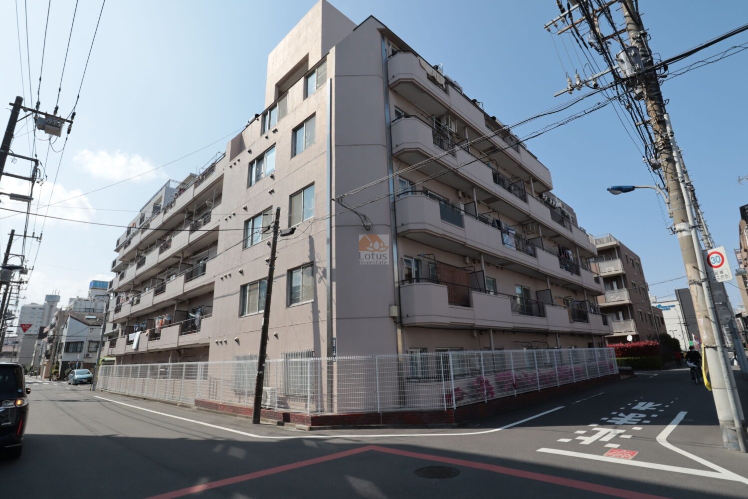 ライオンズマンション荒川遊園の東南角から見た外観です。淡いベージュの外壁が陽を受けて柔らかく映り、下町の住宅街にすっと馴染んでいます。フェンスの足元には花壇が整えられ、日常の中に小さな彩りを感じます。