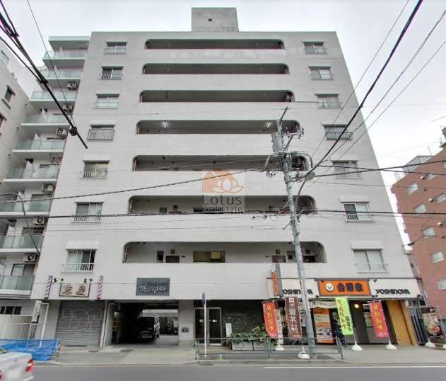 建物は地上8階建の白色外壁で、水平ラインを意識したバルコニーが整然と並んでいます。1階部分には飲食店や店舗が入り、歩道に面して視認性の高いファサードを形成しています。中央部には車両出入口と歩行者用エントランスが確認できます。