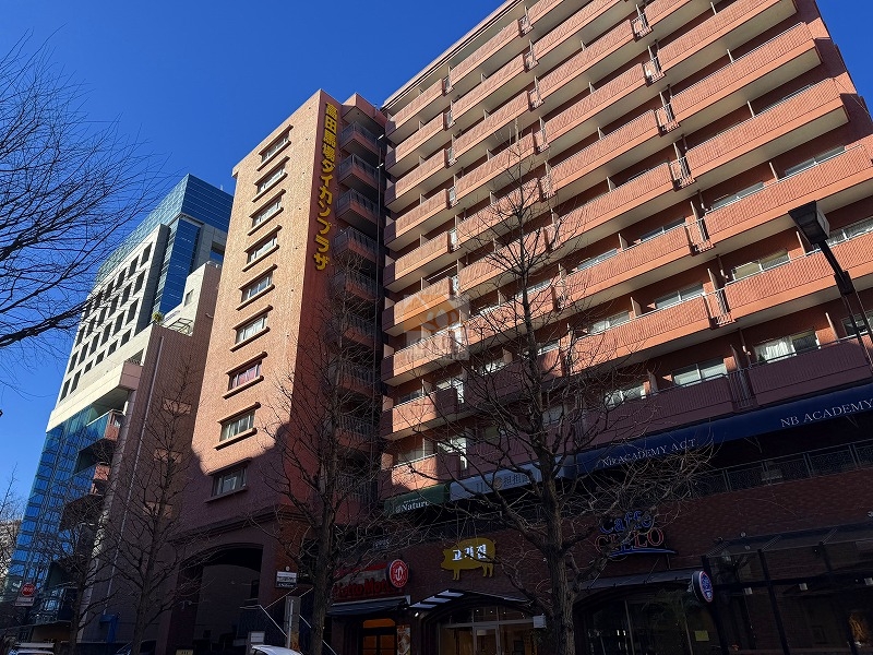 南面バルコニーが連続する中高層の外観が写っており、外壁は煉瓦色系のタイルで統一されています。建物側面に「高田馬場ダイカンプラザ」の縦看板が設置され、下層部には飲食店の袖看板やテナントのファサードが見えます。前面道路沿いには街路樹と街路灯が並び、隣接地にはガラス張りの高層建物が建っています。高田馬場ダイカンプラザは、昭和54年築、11階建てのマンションです。