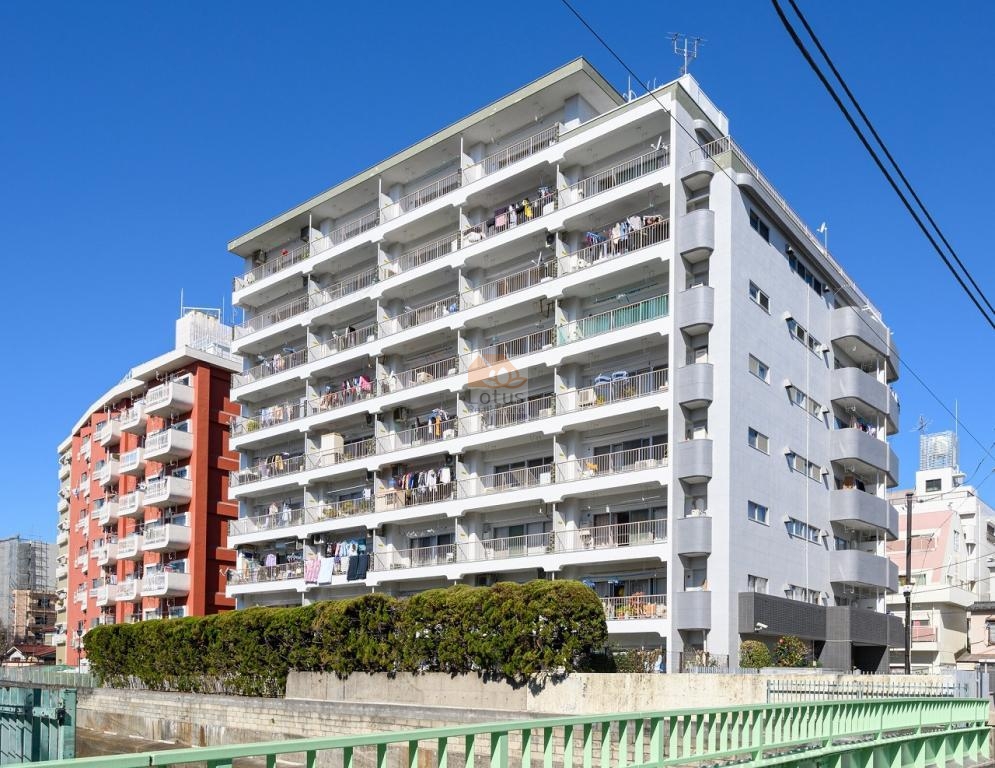 落合マンションの外観（川沿いアングル）。清潔感のある白い外壁に整然と並ぶバルコニーが印象的な中層マンション。南向き中心の住戸設計で、日当たりと風通しにも配慮されている。前面は河川と遊歩道が広がり、開放感ある住環境。落合マンションは、昭和45年築、9階建てのマンションです。
