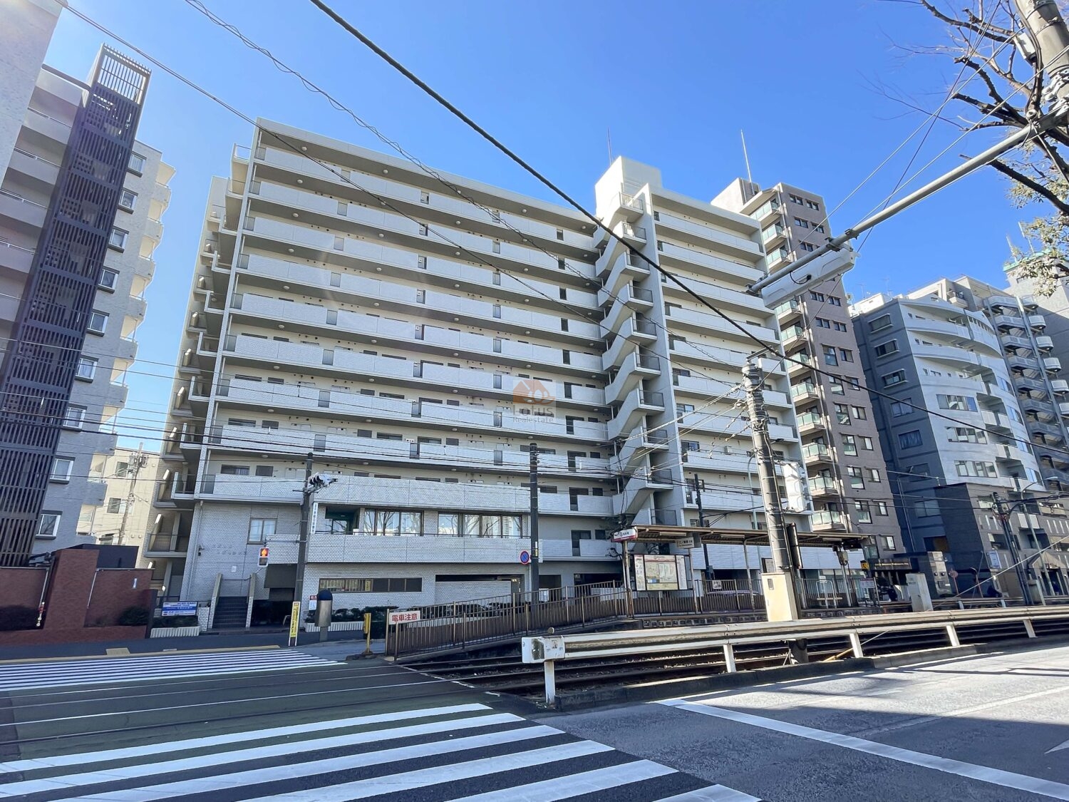 サンファミール西早稲田の外観。白色タイル張りの11階建て住棟で、建物前面には都電荒川線の停留所ホームと軌道、横断歩道、架線設備が写っており、前面道路との取り付きが確認できます。サンファミール西早稲田は、昭和57年築、11階建てのマンションです。新目白通り沿いにあります。