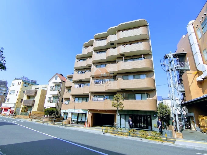 モナーク梅島の建物全景。前面道路に面した6階建てのタイル張り外観が、通りに明るさと統一感を与えている。低層住宅と商業施設が隣接し、街並みに自然に溶け込む。1階部分には店舗スペースが設けられており、生活利便性が高い。モナーク梅島は、平成6年築、6階建てのマンションです。