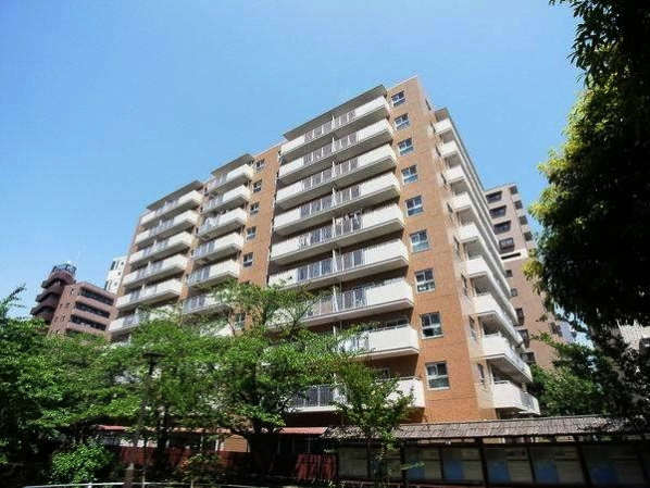 フジイチパークタワーズの外観。レンガ調の外壁を持つ10階建ての建物で、手前には親水公園の樹木があり、緑越しに建物全体を見上げる構図になっています。周囲には中高層マンションが建ち並び、住宅街の一角であることがわかります。フジイチパークタワーズは、昭和56年築、10階建てのマンションです。