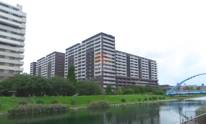 亀戸レジデンスの北東側には旧中川があります。手前に水面、その奥に河川敷の緑地と遊歩道、複数棟が連なる建物が写っています。外壁はダークブラウン系とライトグレー系の配色です。右端にアーチ形のふれいあい橋が見えます。亀戸レジデンスは、平成20年築、17階建てのマンションです。