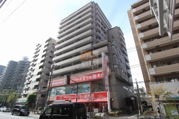 前面道路沿いの全景です。1階は赤色サインのドラッグストアが入居し、ガラス面が連続しています。周囲には中高層の建物が並んでいます。グランドメゾン上池袋は、平成3年築、13階建てのマンションです。