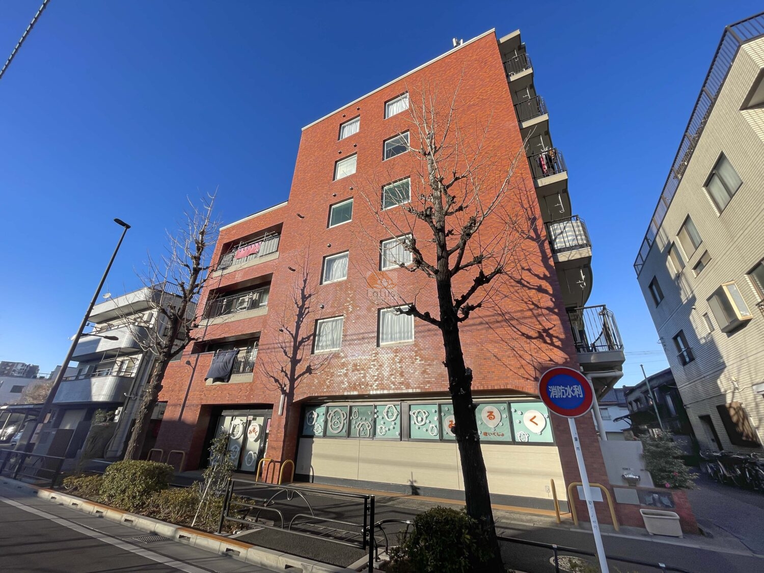 リレント町屋の外観。赤茶色のタイル張りの外観が印象的なマンションです。角地に立地しており、バルコニーには洗濯物や布団が干され、日常の生活感が漂っています。道路沿いには街路樹が植えられ、下町らしい親しみやすい景観が広がっています。リレント町屋は、昭和57年築、8階建てのマンションです。