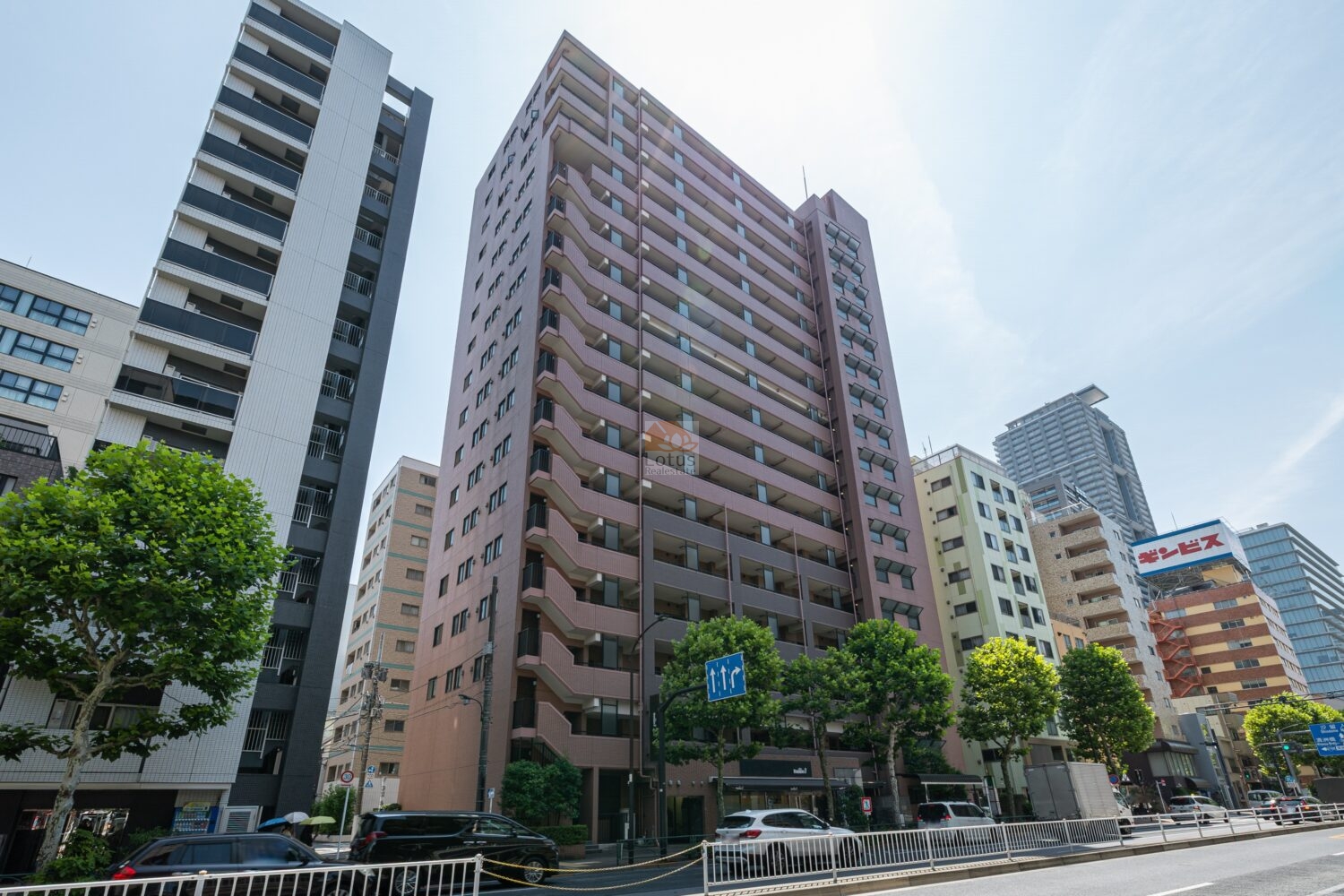 新大橋通りに面した全景です。ピンク系タイルの高層棟が縦に伸び、道路側には折れ線状の外階段と連続するバルコニーが層を作っています。左右には中高層の建物が建ち並んでいます。コスモ日本橋浜町は、平成12年築、17階建てのマンションです。