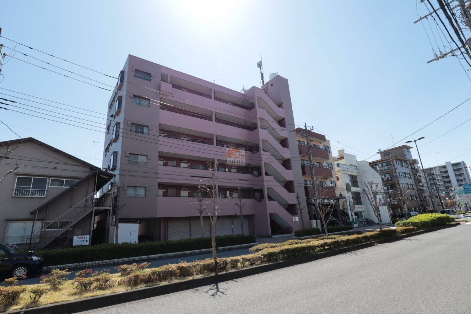 北側の外観は淡いピンク系タイルを基調とした落ち着いた印象で、前面道路との距離が適度に確保されています。建物周囲は住宅街で、歩行空間が広めに取られており人の動線が見えやすい造りです。階段室が外側に開いていて、出入りの様子がつかみやすい構造です。