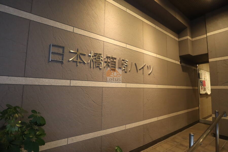 日本橋箱崎ハイツエントランス3｜エントランス壁面には、物件名がスタイリッシュな書体で掲示され、来訪者にマンションの印象を強く残します。照明と緑の配置でシンプルながら洗練された演出がなされています。