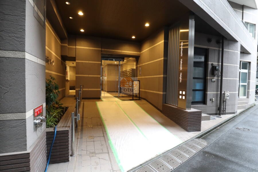 日本橋箱崎ハイツエントランス2｜エントランスアプローチ。天井にダウンライトが並び、雨天時でも明るさを確保できる設計です。奥には集合ポストと自動ドアがあり、利便性と安心感を兼ね備えています。