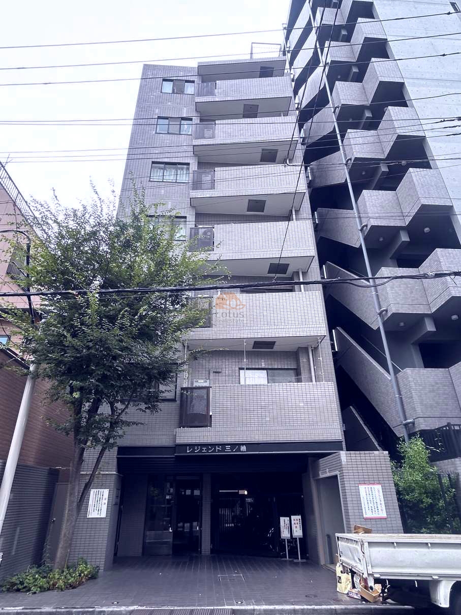 グレー系のタイル張りの落ち着いた外観の建物で、エントランス上部には「レジェンド三ノ輪」の文字が見えます。建物の手前には緑の木が植えられています。エントランス部分は車路のような造りになっていて、奥にマンションの入り口が見える構造になっていますね。レジェンド三ノ輪は、平成2年築、8階建てのマンションです。