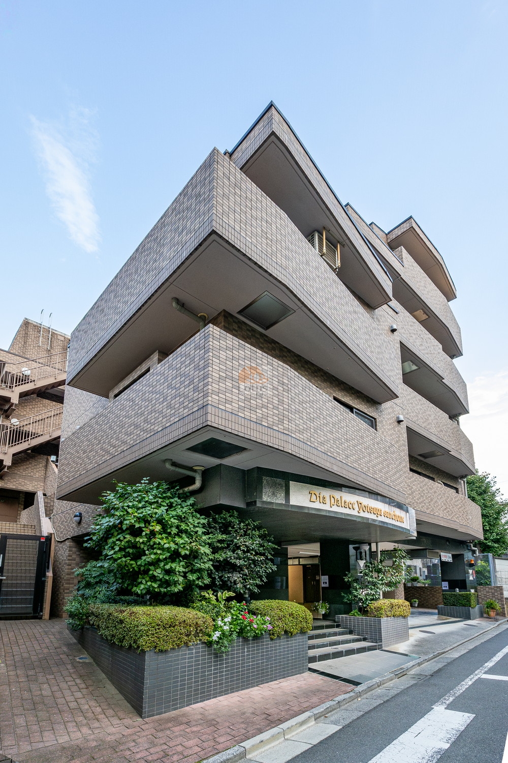 建物の外観を見上げるアングルで撮影されており、タイル貼りの低層マンションが確認できます。バルコニーが段状に配置され、街路に面した敷地には植栽が整えられています。ダイアパレス四谷三丁目は、平成10年築、4階建てのマンションです。