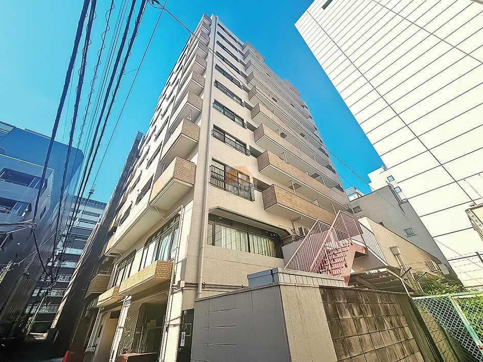 路地側から見上げた外観で、11階建ての住棟が縦に伸び、バルコニーが交互に張り出しています。低層部は連続ガラスとタイル基壇で構成され、右手には外部階段と擁壁、上空には電線が通っています。中銀音羽マンシオンは、昭和55年築、11階建てのマンションです。