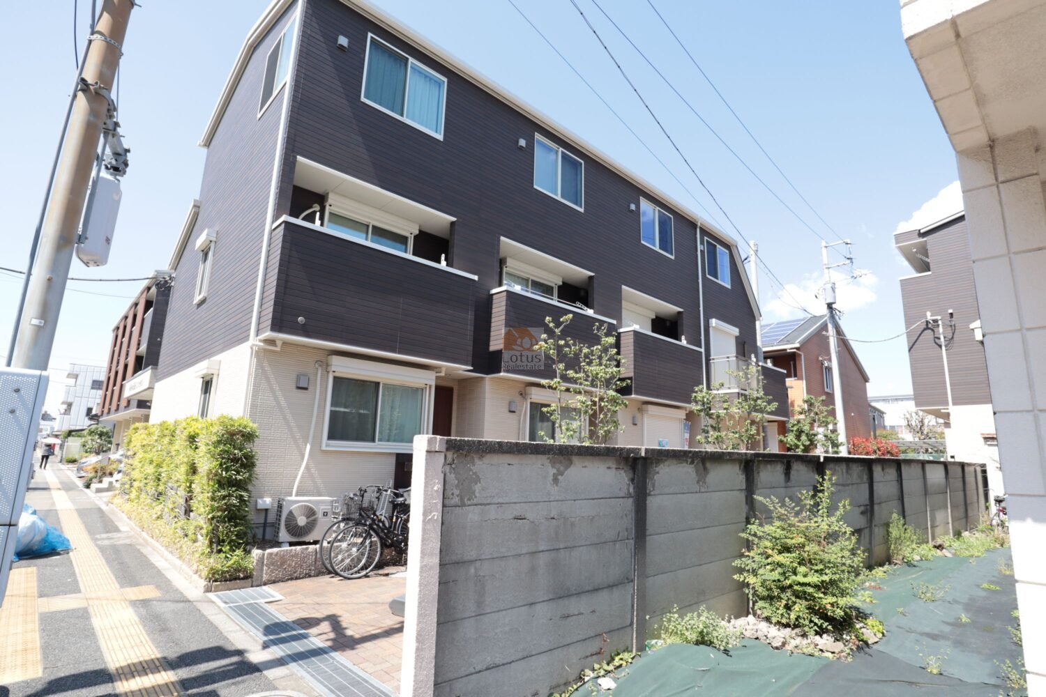 スマｅタウン足立竹ノ塚の外観全景が写っていて、ダークブラウンの外壁と明るいベージュの低層部が組み合わさった3階建てのタウンハウスが道路沿いに並んでいます。前面には植栽とインターロッキング舗装のアプローチが続き、歩道と建物の間にほどよい距離感をつくっている様子が見えてきます。前面道路の車線や電柱も写り込んでいて、竹ノ塚エリアの生活道路に面した日常的な街並みの中で暮らしているイメージが湧く構図です。