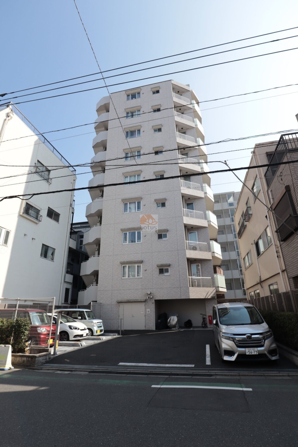 プレジール尾久ステーションマークスの外観（北側）を正面から見上げています。前面に平置き駐車スペースとスロープ、奥にピロティの出入口が見えて、道路から車や自転車でそのままアプローチできる動線になっています。外壁は淡色タイルとガラス手すりが連なり、下町の静かな路地に溶け込みながら清潔感を出しています。