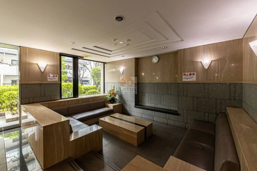 ライオンズガーデン錦糸町ロビー1｜ライオンズガーデン錦糸町のロビーラウンジ。大型の窓面から植栽が見える空間に、L字型のソファとテーブルが配置されている。照明付きの壁面パネルと時計が設置された落ち着いた空間。