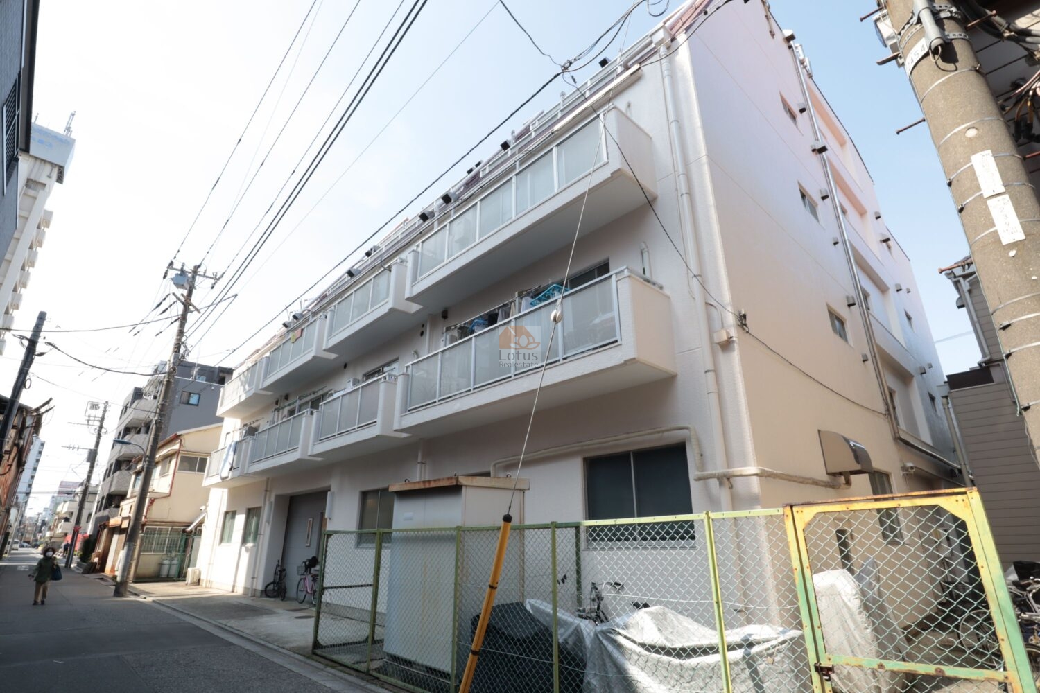 コーポ尾久の南西側の外観です。細い生活道路に面して、連続するガラス手すりのバルコニー、電柱と電線が頭上を走り、建物脇には自転車とフェンスで囲われたスペースが見えてきます。