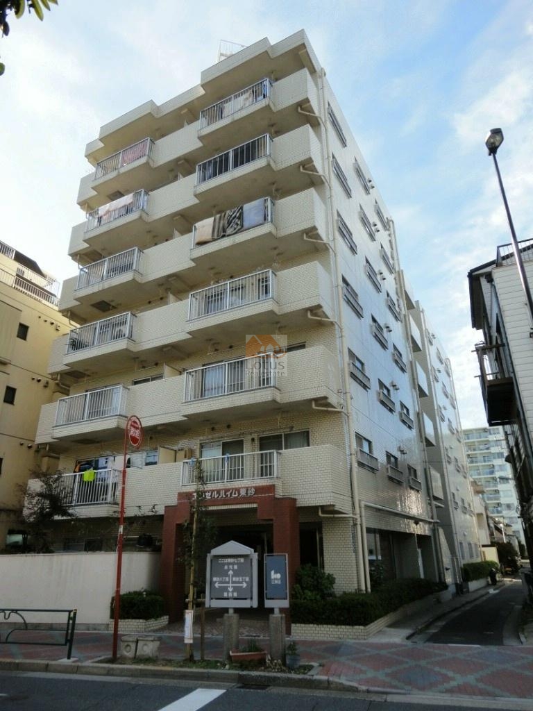 エンゼルハイム東砂の外観。白いタイル貼りの外壁が特徴的な7階建てのマンションで、各住戸にはバルコニーが設けられています。低層部には植栽が配置され、通りに面して赤いポールや歩道が整備されているのが見えます。エンゼルハイム東砂は、平成元年築、7階建てのマンションです。