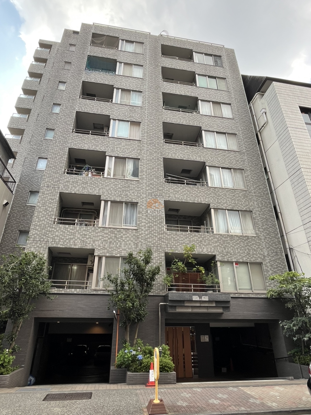 グレー系タイルの外壁に、開口の深いバルコニー、1階は左右に車路の開口があり、中央に木目調の出入口が見えます。歩道側には植栽のあるプランターと樹木があります。ユニオンパレス御徒町は、平成13年築、12階建てのマンションです。