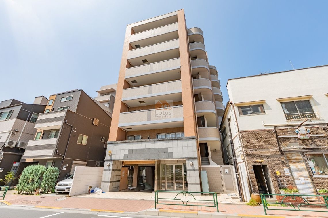 正面から見た外観です。オレンジ色の縦ラインと白いバルコニーが組み合わさったデザインで、8階建ての建物がすっきりと伸びています。左右には低層の建物が並び、街並みの中で存在感を見せています。ロータリーパレス田端は、平成15年築、8階建てのマンションです。