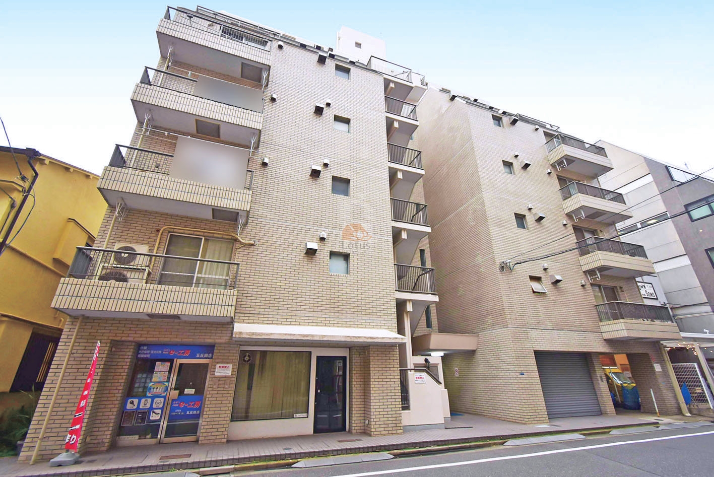 エクレール五反田の外観（通り沿い全景）。ベージュ系タイル貼りの建物で、低層階には店舗区画とシャッター付きの駐車スペースを併設。都市型ヴィンテージマンションとしての実用性と存在感を備えています。エクレール五反田は、昭和55年築、8階建てのマンションです。