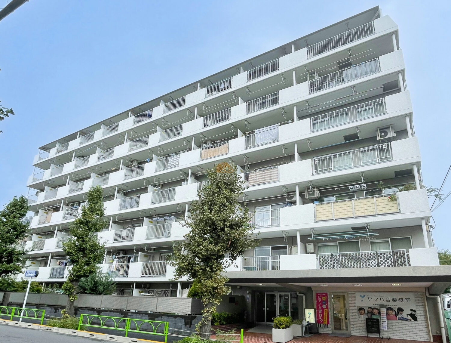 千歳船橋ビューグリーンの外観。白を基調とした7階建の外壁に、各住戸のバルコニーが整然と並ぶデザイン。前面の緑と歩道が、街並みに調和した穏やかな住宅環境を印象づけます。千歳船橋ビューグリーンは、昭和53年築、７階建てのマンションです。