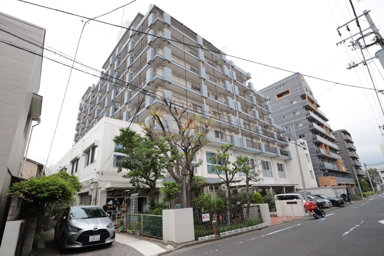 パシフィック第2門前仲町マンションの外観で、白い外壁にグレー系のバルコニーが連続し、角地側に植栽と低い塀が見えます。前面道路は幅があり、車の出入りがしやすい距離感です。電線が視界に入るぶん、生活圏のリアルさも伝わる構図です。