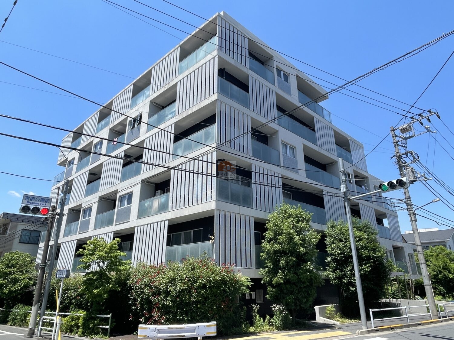 交差点の角地に建つ中層レジデンスです。ファサードは縦ルーバーとガラス手すりのバルコニーが交互に並び、水平スラブが段ごとに見えます。街路側には植栽があります。ウィンシティ成増は、平成18年築、6階建てのマンションです。