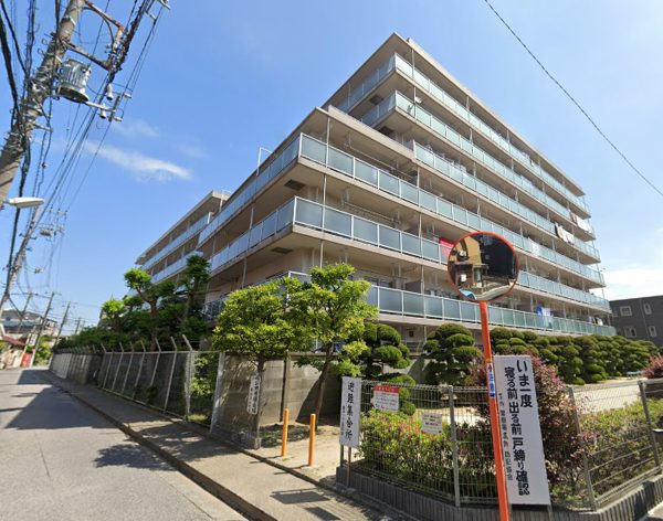 角地の外観です。敷地周囲は金網フェンスと植栽で囲われ、刈り込みされた低木が並びます。道路にはカーブミラー、電柱と架線、注意看板が立っています。奥戸ハウスは、昭和55年築、7階建てのマンションです。