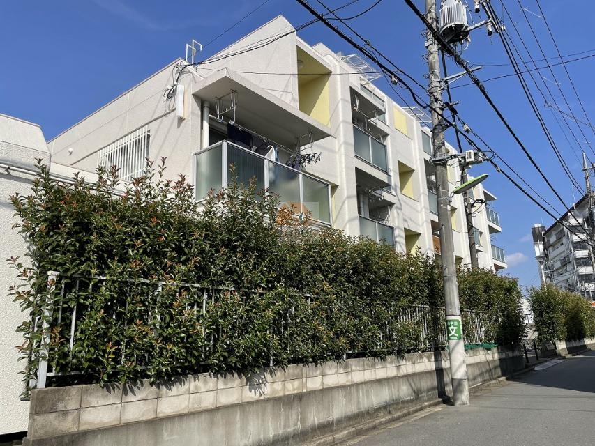 白い外壁に、奥まったバルコニーと半透明のガラス手すりが連なり、各住戸の開口部付近には淡いイエローのアクセントが入り、道路側は生垣とコンクリート塀で連続しています。電柱や配線が道路上に走り、建物は角度を付けて配置されています。上用賀マンションは、昭和51年築、4階建てのマンションです。