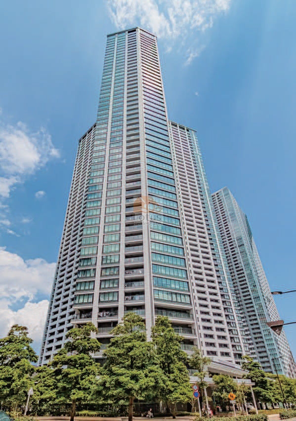 勝どきザ・タワーの外観。地上53階建ての外観が青空に向かってそびえ立ち、全面に広がるガラスの窓面が都市の光を映し出しています。足元には整然と植栽された緑が広がり、巨大な建物の存在感と都市の中の潤いが同居しています。勝どきザ・タワーは、平成28年築、53階建てのタワーマンションです。