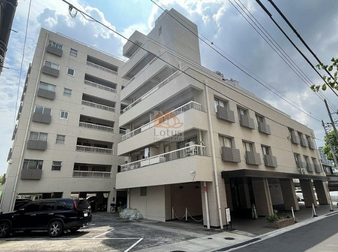 白金武蔵野コーポラスの外観。建物はベージュ系の外壁で統一され、7階建ての中層構造です。正面には庇のあるエントランスと、車寄せに利用できるピロティ形式の駐車スペースが設けられています。建物の前面には舗装された駐車場があります。空は雲が多めで、建物の立体感が際立っています。白金武蔵野コーポラスは、昭和45年築、7階建てのマンションです。