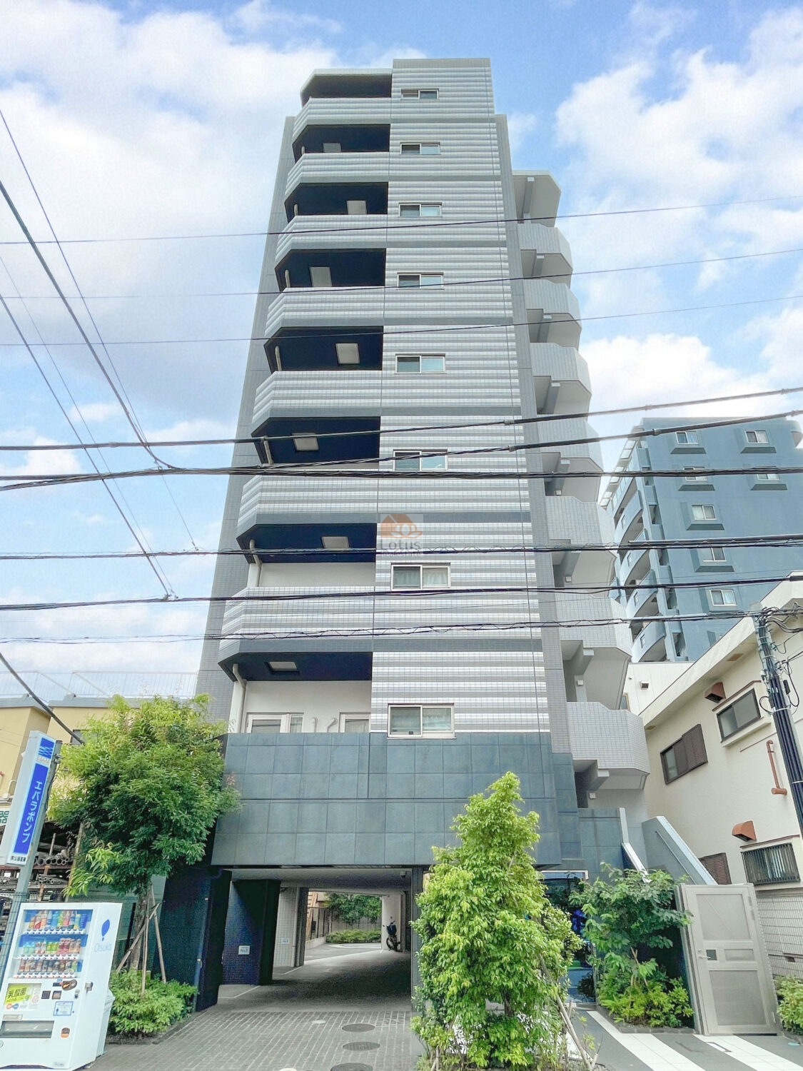 建物はグレー系のタイル貼りで統一された9階建の外観です。縦のラインが強調されたデザインで、1階部分はピロティ構造で、奥に駐輪スペースが見えます。正面には植栽と自動販売機が設置され、通り沿いにも緑が配置されています。ヴィルヴィオ葛西は、平成29年築、9階建てのマンションです。