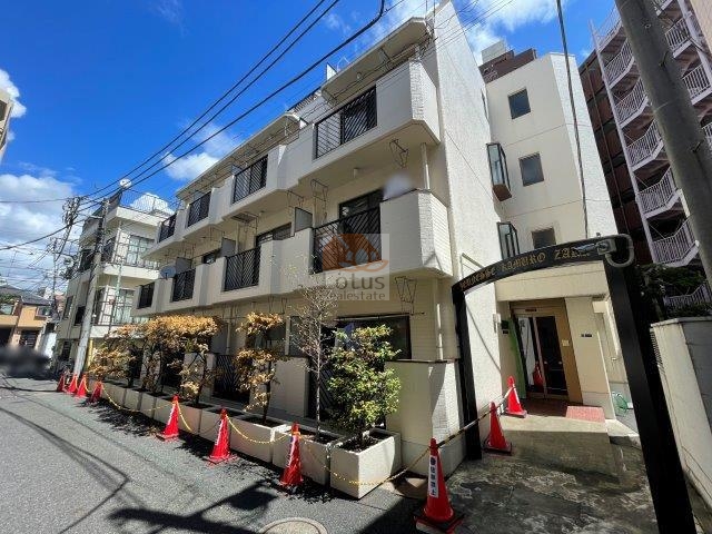 ジュネスかむろ坂の外観：白い外壁と黒の格子バルコニーが整然と並ぶ4階建ての低層レジデンス。通り沿いには緑を取り入れた植栽が設けられ、都市生活にささやかな潤いを添えている。小規模ながらも丁寧に手入れされた建物正面が印象的。ジュネスかむろ坂は、昭和58年築、4階建てのマンションです。