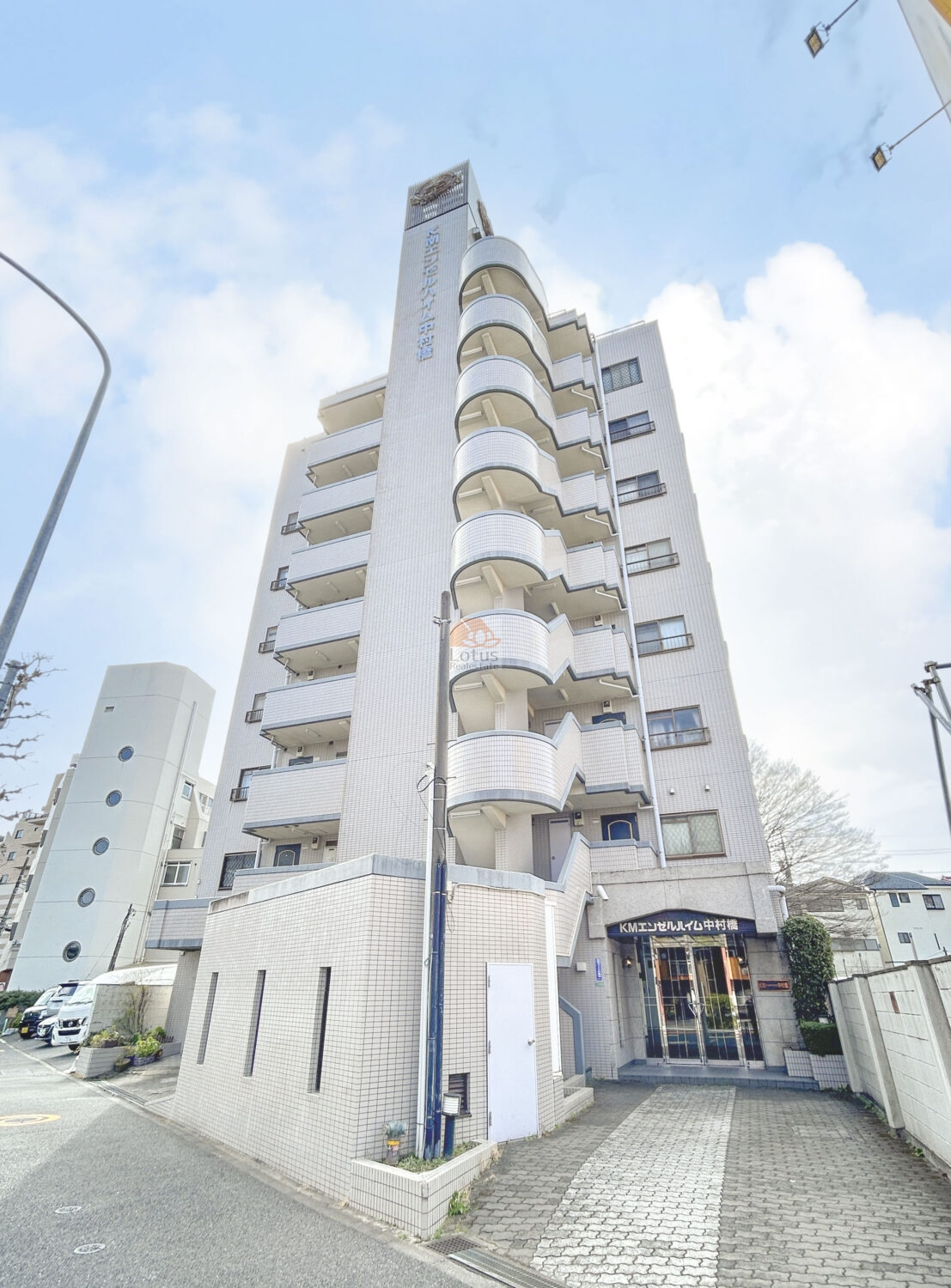 白いタイル張りのKMエンゼルハイム中村橋を見上げる構図です。角地に建ち、道路側には丸みのあるバルコニーが縦に連なっています。右手のガラス扉のエントランス上には「KMエンゼルハイム中村橋」のサインがあり、前面はブロック舗装のアプローチと低い塀が続きます。街路灯と電柱、隣接する建物、背後の樹木、青空と雲までが写っています。KMエンゼルハイム中村橋は、平成4年築、8階建てのマンションです。