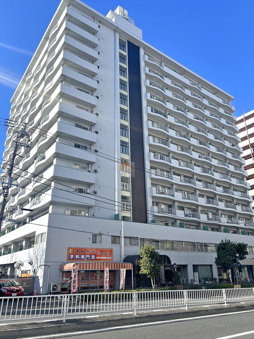 クレールメゾン品川の外観。白系外壁の高層マンションで、中央の縦アクセントパネル、屋上設備が見え、1階はテナント区画（学科専門塾の看板とオーニング）とガラス面が並び、前面は歩道・ガードレール・植栽が整い、視認性が高く来客案内に使いやすい。クレールメゾン品川は、昭和50年築、14階建てのマンションです。