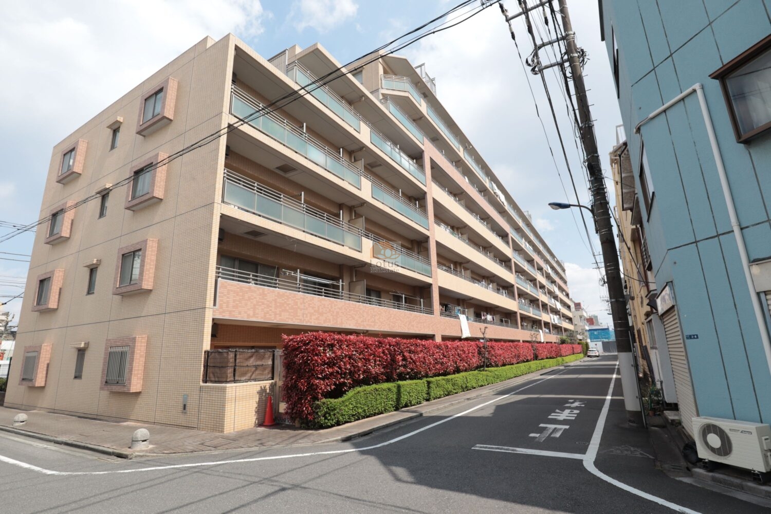 リーデンススクエア滝野川の外観です。南西向きのバルコニーが明るい街並みを映しています。植栽の赤い生け垣が通りを彩り、日常の風景にあたたかさを添えています。